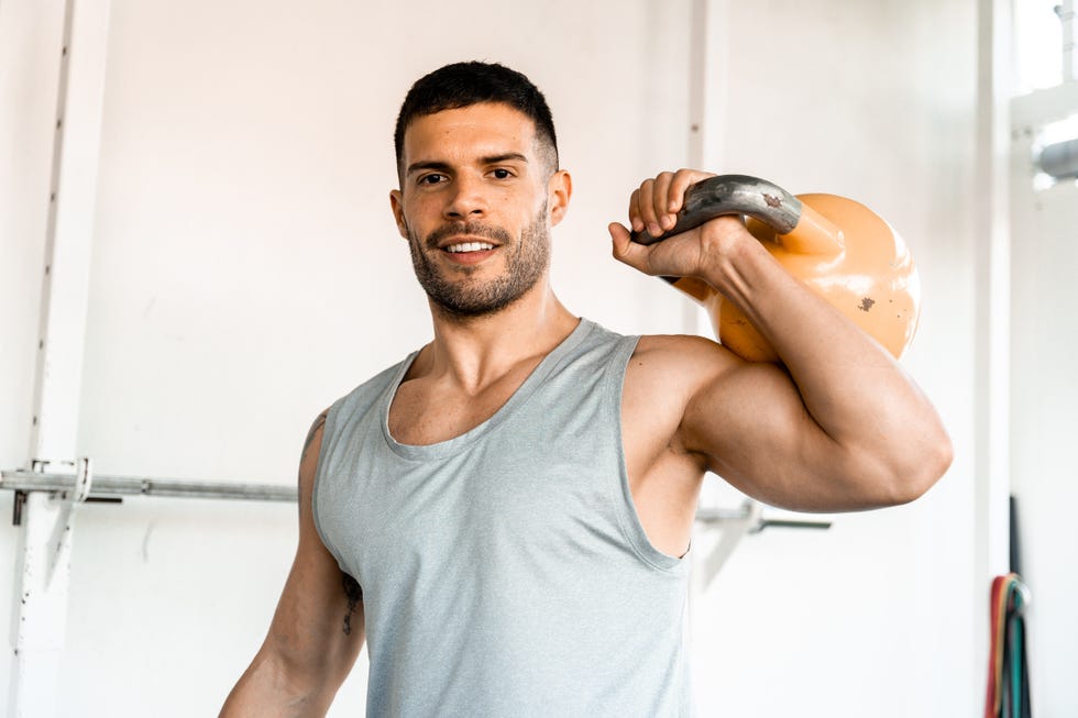Man smiling at the camera while holding kettlebell over his shoulder. man smiling at the camera while holding kettlebell over his shoulder male athlete holding kettlebell doing gym workout in gym