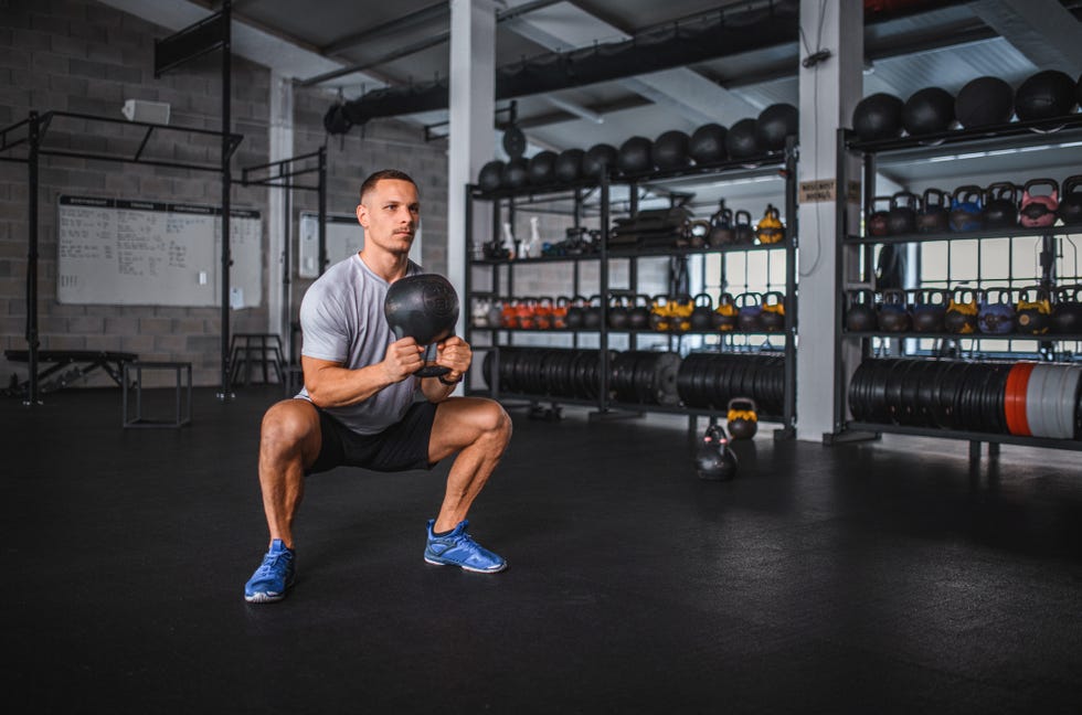 Caucasian Male Athlete Doing Kettlebell Sumo Squat in Gym full length front view of sportsman in early 30s working out with kettlebell in spacious cross training facility