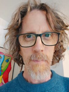 Joel Merriner is a bespectacled man of European descent who has more than a little grey in his red beard. He's very close to the camera, mostly blocking the wall behind himself, but there is a corner of a picture showing on the wall and I cannot for the life of me tell if it's a fancy depiction of the tragedy mask or a map with a waterfall running through it.